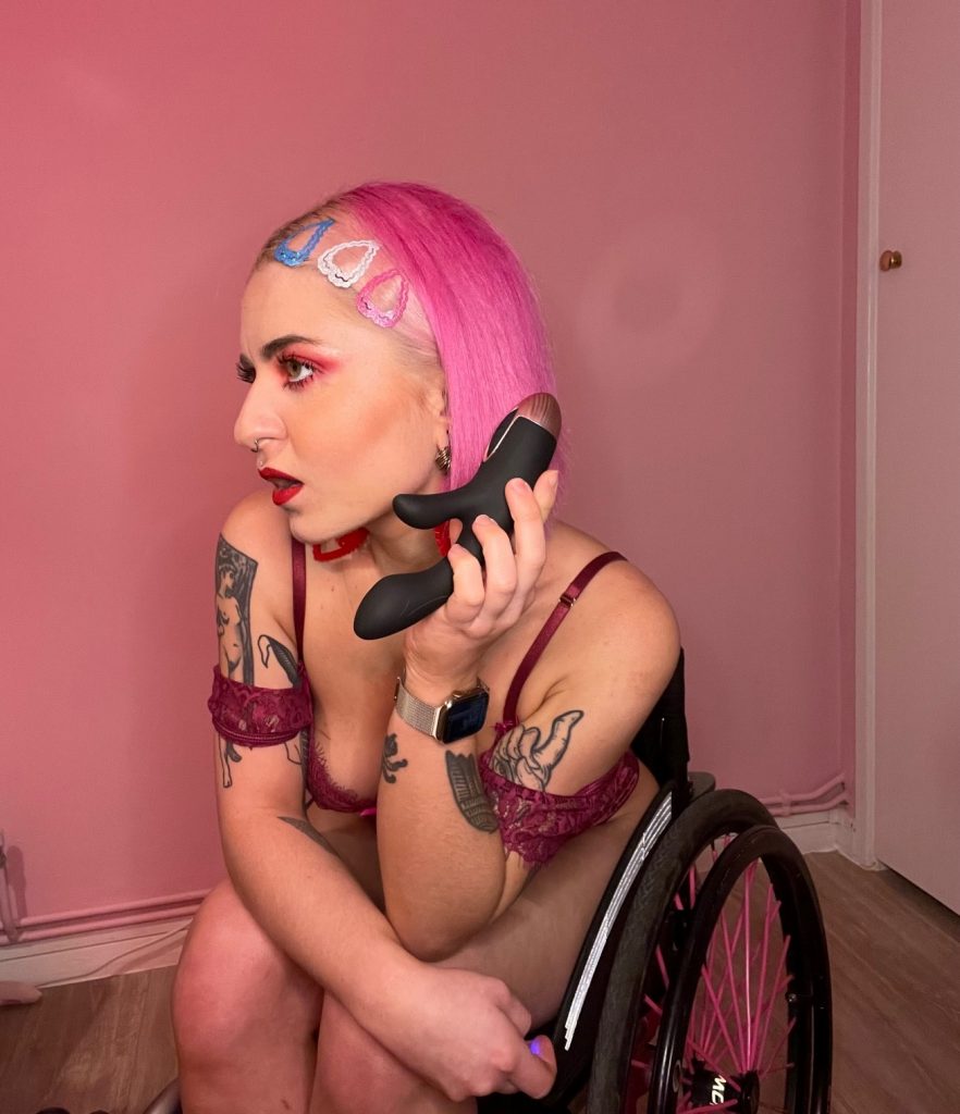 Bondara Sex Toys Blog - Sex & Disability: An Interview with Disabled_Eliza & TheChronicIconic - Jess sitting in her chair smiling, wearing the Bondara Coquette Velvet Garter Teddy, holding the Bondara Warming Rabbit Vibrator like a phone